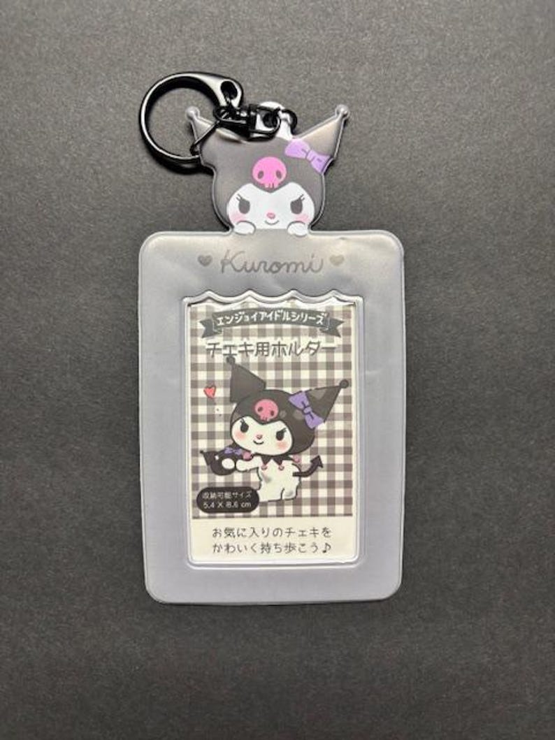 May include: A gray Kuromi-themed keychain with a cartoon character design. The keychain features a clear plastic holder with a printed image of Kuromi and Japanese text. The holder is designed to hold a small card, measuring 2.1 x 3.4 inches.