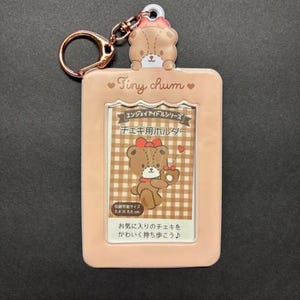 May include: A light pink Tiny Chum photo holder with a bear design, a gold-colored keychain, and Japanese text. The holder features a bear illustration holding a cherry and a checkered background. The text reads "Tiny Chum".