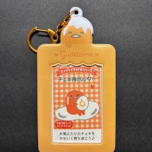 May include: A yellow Gudetama keychain with a gold-colored keyring. The keychain features a rectangular design with a clear window displaying a cartoon image of Gudetama. The text "Gudetama" is printed on the keychain.