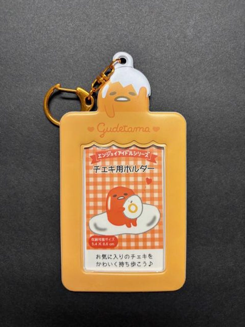 May include: A yellow Gudetama keychain with a gold-colored keyring. The keychain features a rectangular design with a clear window displaying a cartoon image of Gudetama. The text "Gudetama" is printed on the keychain.