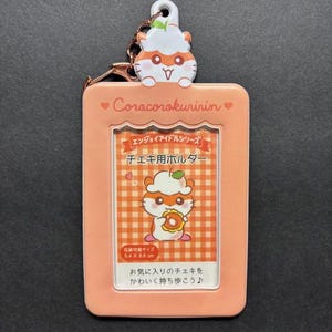 May include: A peach-colored photo holder with a cartoon hamster design. The holder has a clear window for a photo and a matching hamster charm. Text on the holder includes "Corocorokuririn" and Japanese text. The holder is 5.4 cm x 8.6 cm.