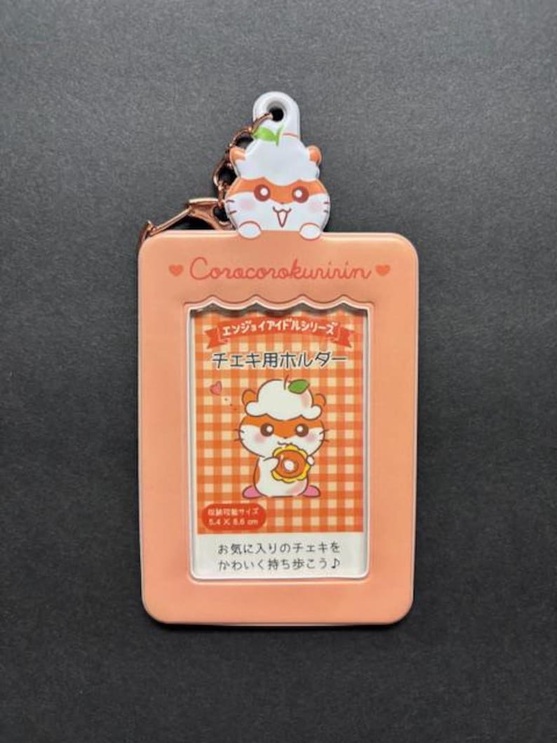 May include: A peach-colored photo holder with a cartoon hamster design. The holder has a clear window for a photo and a matching hamster charm. Text on the holder includes "Corocorokuririn" and Japanese text. The holder is 5.4 cm x 8.6 cm.