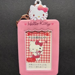 May include: A pink Hello Kitty photo holder with a metal clasp. The holder features a Hello Kitty character at the top and a rectangular frame with a picture of Hello Kitty. The text on the holder says "Hello Kitty" and includes Japanese text.