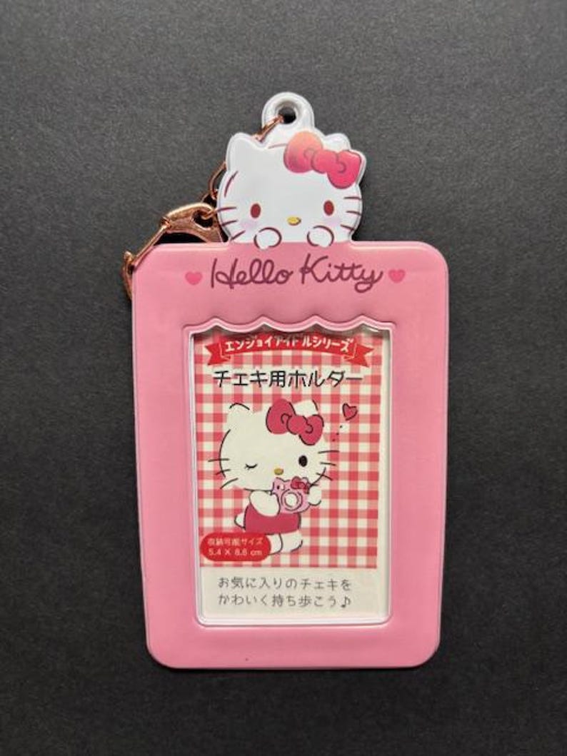 May include: A pink Hello Kitty photo holder with a metal clasp. The holder features a Hello Kitty character at the top and a rectangular frame with a picture of Hello Kitty. The text on the holder says "Hello Kitty" and includes Japanese text.