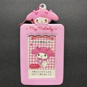 May include: A pink My Melody ID holder with a matching charm. The holder features a picture of My Melody and text in Japanese. The holder is designed to hold a 5.4 x 8.6 cm photo.