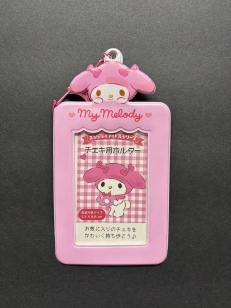 May include: A pink My Melody ID holder with a matching charm. The holder features a picture of My Melody and text in Japanese. The holder is designed to hold a 5.4 x 8.6 cm photo.