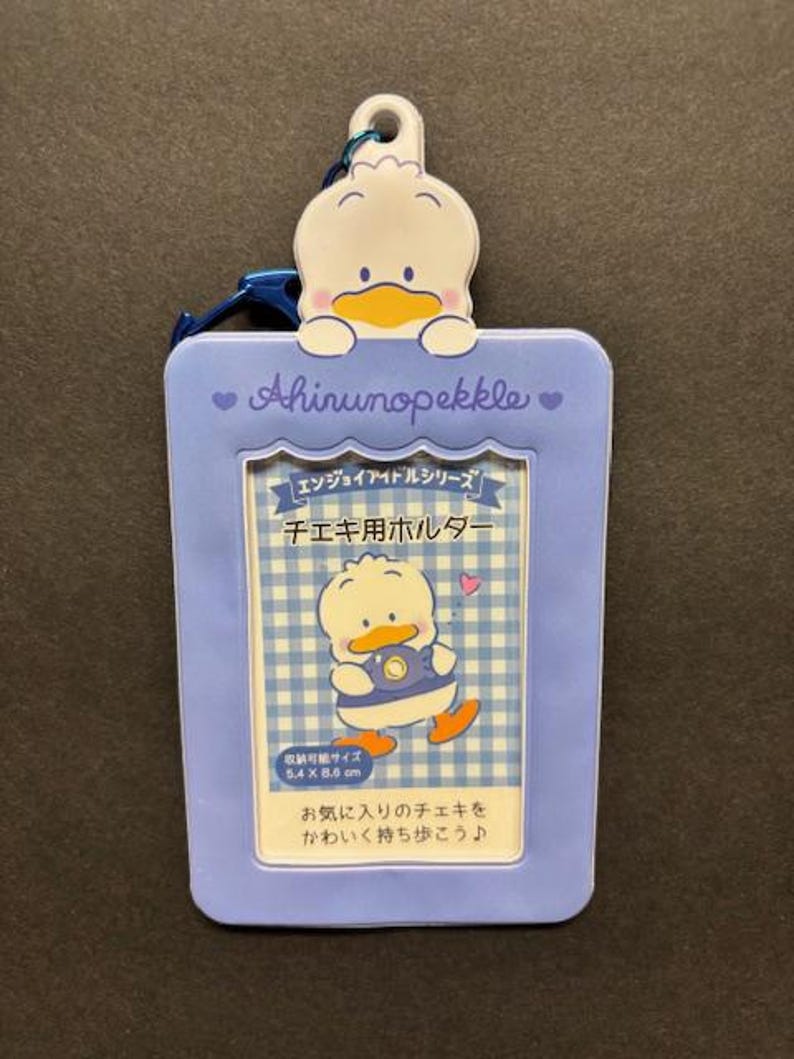 May include: A light blue photo holder featuring a cartoon duck character. The holder has a rectangular shape with rounded edges and a small duck-shaped charm at the top. The text "Ahinunopekkle" is printed on the holder.