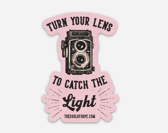 Puede incluir: Pegatina rosa con una ilustración de cámara vintage y el texto "Gira tu lente para capturar la luz" con el sitio web "thesoulofhope.com" debajo.