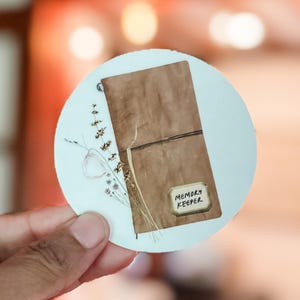 Puede incluir: Una pegatina redonda con un diario marrón con cierre de cuerda y las palabras "MEMORY KEEPER" en una etiqueta. La pegatina tiene un estilo de acuarela con ramitas de flores secas y vegetación.