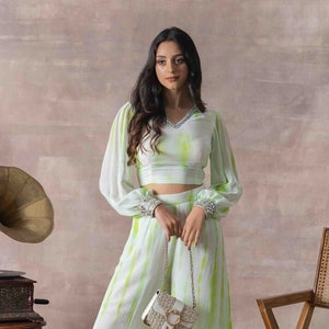 May include: A white and green tie-dye crop top and wide-leg pants set with a white and gold chain strap purse. The top has a V-neck and long sleeves with beaded embellishments on the cuffs. The pants have a wide leg and a tie-dye pattern.