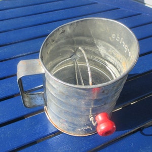 Puede incluir: Un tamiz de harina vintage de metal con una manivela roja. El tamiz tiene una malla de alambre y un asa en el lateral. El tamiz está marcado con "Ebira" en el lateral.