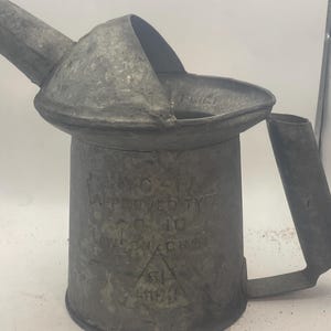 May include: A vintage metal oil can with a spout and handle. The can is a dull gray color and has text that reads "APPROVED TYPE" and "10". The can has a triangular logo with the letters "SL" inside.