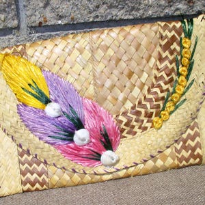 Könnte beinhalten: Eine geflochtene Strohtasche mit einem braunen und beigefarbenen Chevron-Muster. Die Tasche ist mit lila, rosa und gelben Blumen und weißen Muscheln verziert.