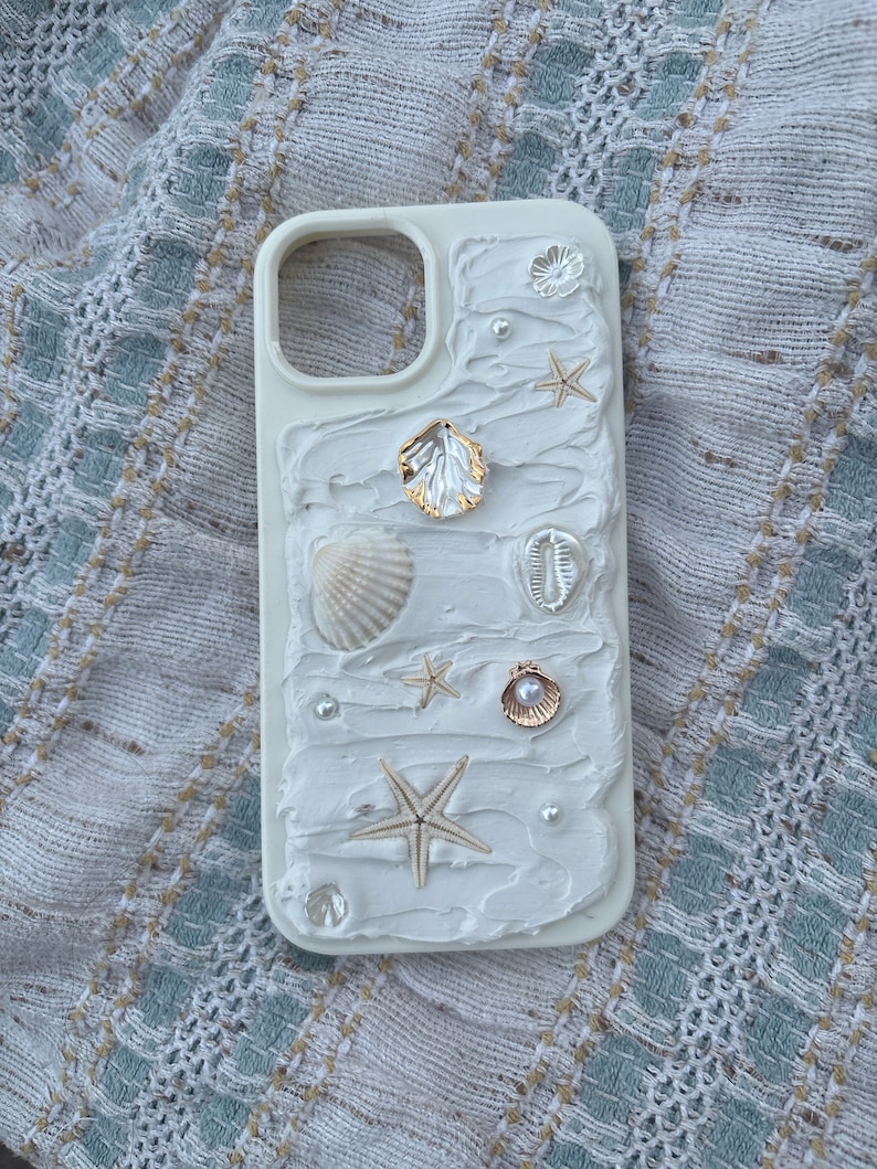 May include: White phone case with a textured, stucco-like design. Adorned with seashells, starfish, pearls, and a gold-colored shell. The case has a cutout for the camera and buttons.