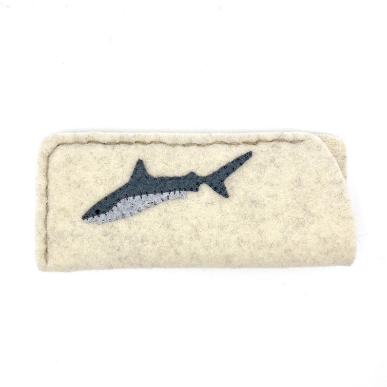 Op de afbeelding: Een rechthoekig etui van beige vilt met een donkergrijze haaienapplicatie op de voorzijde. Het etui is voorzien van zichtbare donkere stiksels langs de randen en op het haaienontwerp, wat zorgt voor een getextureerd en handgemaakt uiterlijk.