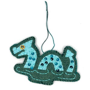 Può includere: Un ornamento fatto a mano in feltro a forma di mostro marino. Il mostro marino è verde acqua con dettagli blu e paillettes. Ha un occhio giallo e un cordino verde per appenderlo. L'ornamento è lungo circa 10 cm.