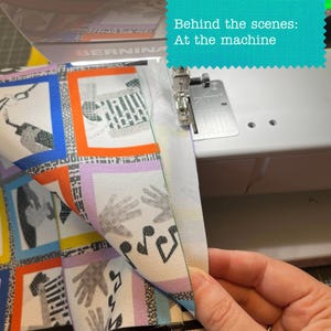 Op de afbeelding: Een witte Bernina naaimachine stikt stof met een kleurrijk ruitpatroon van handen en muzieknoten. De stof heeft oranje, blauwe, gele en paarse randen. Tekst op een groenblauwe achtergrond luidt: Behind the scenes: At the machine.