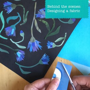 Op de afbeelding: Een zwart oppervlak toont uitgesneden papieren korenbloemen in blauw- en paarstinten met groene stengels en bladeren. Een turquoise rechthoekig label bevat de witte tekst "Behind the scenes: Designing a fabric" terwijl een schaar papier knipt.