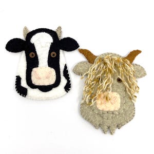 May include: Two felt cow ornaments. One is black and white with a pink nose and brown eyes. The other is grey with a tan nose, brown eyes, and tan yarn hair. Both have small horns.