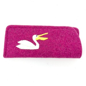 May include: A rectangular magenta felt case featuring an appliqued white pelican with a yellow beak. The pelican is depicted in profile with its wings folded against its body. The case has a simple, flat construction with visible felt texture.