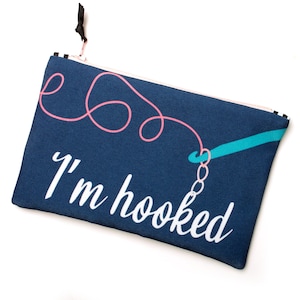 Puede incluir: Un estuche rectangular de color azul marino con cremallera que presenta un gr&aacute;fico de un ganchillo turquesa y lana rosa formando bucles. El texto blanco "I'm hooked" est&aacute; impreso en cursiva en la parte inferior de la tela.