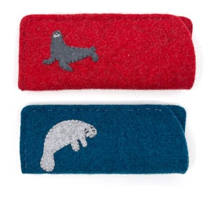 Op de afbeelding: Twee rechthoekige vilten etuis met gestikte randen. Het bovenste etui is felrood en voorzien van een donkergrijze zeehond-applicatie. Het onderste etui is diepblauw en voorzien van een lichtgrijze zeekoe-applicatie.