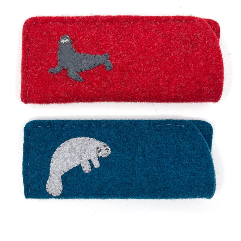 Op de afbeelding: Twee rechthoekige vilten etuis met gestikte randen. Het bovenste etui is felrood en voorzien van een donkergrijze zeehond-applicatie. Het onderste etui is diepblauw en voorzien van een lichtgrijze zeekoe-applicatie.