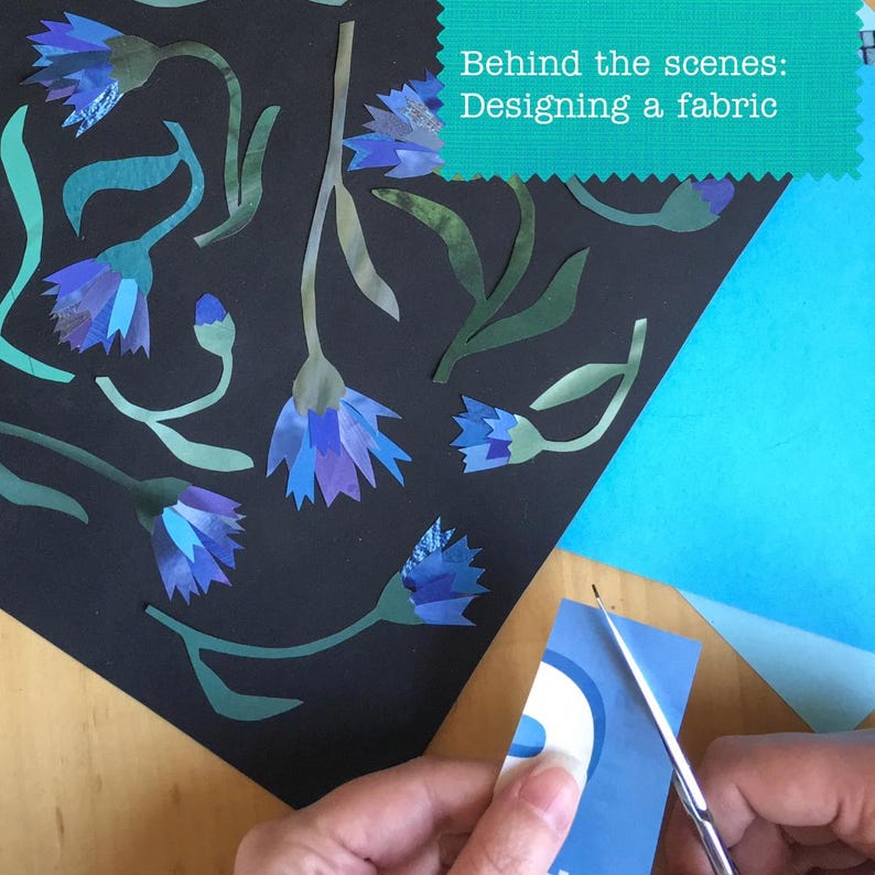May include: A black surface features cut-out paper cornflowers in shades of blue and purple with green stems and leaves. A teal rectangular label displays the text "Behind the scenes: Designing a fabric" in white, while scissors cut blue paper.
