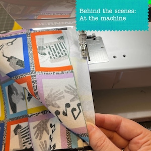 Puede incluir: Una m&aacute;quina de coser Bernina blanca cosiendo una tela con un patr&oacute;n de cuadr&iacute;cula colorido que presenta ilustraciones de manos y notas musicales. La tela tiene bordes naranjas, azules y amarillos. El texto dice: Behind the scenes: At the machine.