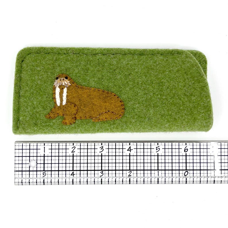 Op de afbeelding: Een rechthoekig groen vilten etui met een geborduurde bruine walrus met witte slagtanden. Een metalen liniaal eronder toont een lengte van ongeveer 16,5 cm en een hoogte van ongeveer 6,5 cm.