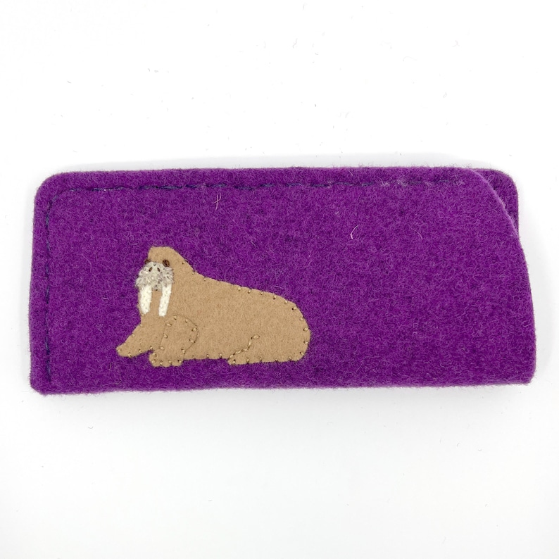 Op de afbeelding: Een rechthoekig etui van paars vilt met een beige walrus-applicatie aan de linkerkant. De walrus is in profiel afgebeeld met witte slagtanden en gedetailleerde stiksels langs het lichaam. Het etui heeft een eenvoudige, platte constructie.