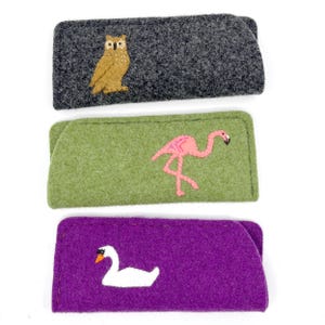 May include: Three rectangular felt cases for glasses. The top case is dark gray with a brown owl embroidery. The middle case is light green with a pink flamingo embroidery. The bottom case is purple with a white swan embroidery on the front.