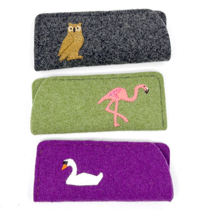 May include: Three rectangular felt cases for glasses. The top case is dark gray with a brown owl embroidery. The middle case is light green with a pink flamingo embroidery. The bottom case is purple with a white swan embroidery on the front.