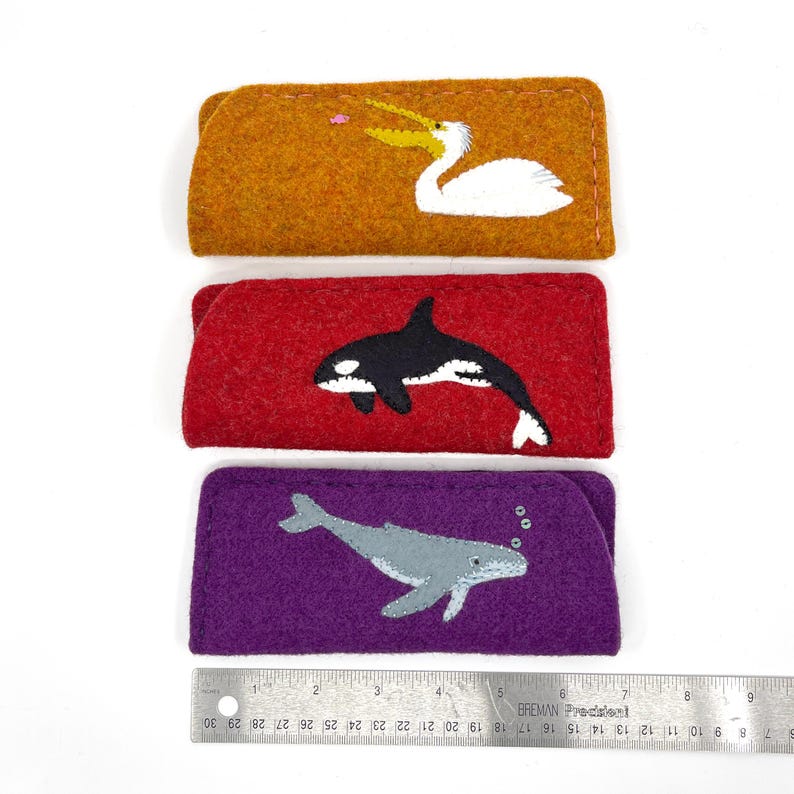 Op de afbeelding: Drie rechthoekige vilten etuis van ongeveer 18 cm lang. Het bovenste etui is mosterdgeel met een witte pelikaan, het middelste is rood met een zwart-witte orka en het onderste is paars met een grijze walvis en kleine luchtbelletjes.