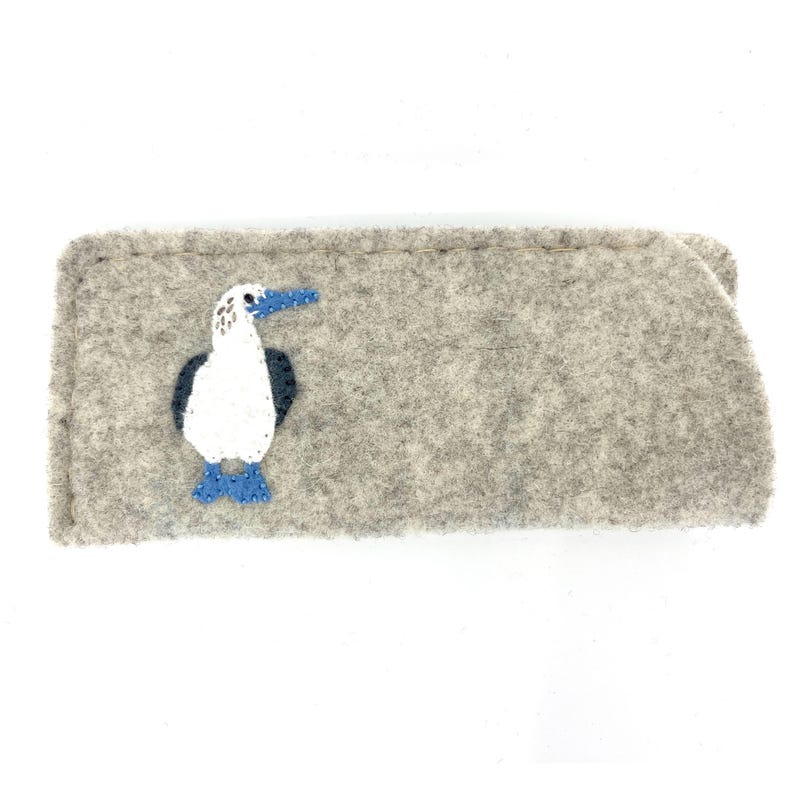 May include: A rectangular light gray felt case featuring an embroidered blue-footed booby bird. The bird has a white body, dark gray wings, and bright blue feet and beak. The case is constructed with visible stitching along the edges.