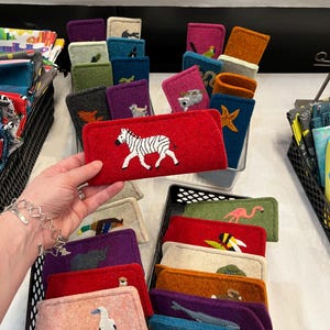 Op de afbeelding: Een collectie rechthoekige vilten etuis in diverse kleuren, elk voorzien van een uniek geborduurd diermotief. De etuis zijn georganiseerd in zwarte plastic manden, waarbij &eacute;&eacute;n rood etui met een wit-zwarte zebra op de voorgrond wordt vastgehouden.