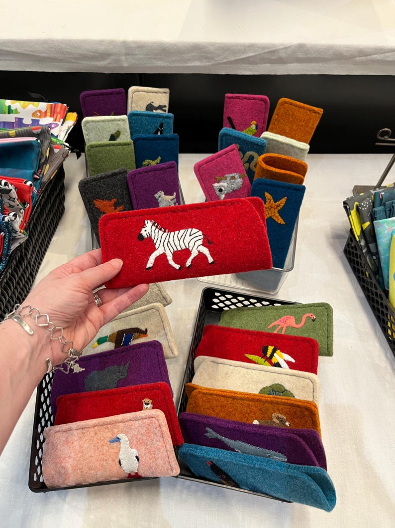 Op de afbeelding: Een collectie rechthoekige vilten etuis in diverse kleuren, elk voorzien van een uniek geborduurd diermotief. De etuis zijn georganiseerd in zwarte plastic manden, waarbij &eacute;&eacute;n rood etui met een wit-zwarte zebra op de voorgrond wordt vastgehouden.