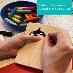 May include: A beige felt square featuring a black and white orca embroidery design, being stitched with black thread. A blue bowl filled with various colorful sewing thread spools sits nearby. Text in the image reads: Behind the scenes: At work in the studio.