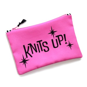 May include: Pink zippered pouch with black stars and the text "KNITS UP!" printed on it.