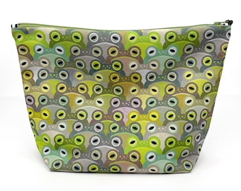 Puede incluir: Un estuche trapezoidal con cremallera que presenta un patrón repetitivo de caras de rana estilizadas en varios tonos de verde lima, oliva, salvia y gris. El diseño consiste en cabezas de rana superpuestas con ojos prominentes y coloridos.