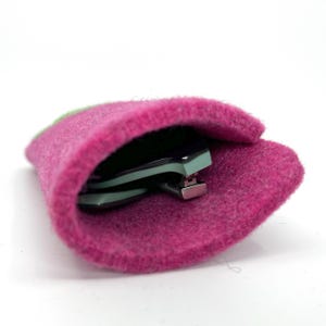 Op de afbeelding: Een fel magenta viltzakje met een zacht, getextureerd oppervlak. Binnenin de opening van het zakje is een opgevouwen bril met licht muntgroene monturen en zilverkleurige metalen scharnieren gedeeltelijk zichtbaar in de hoes geplaatst.
