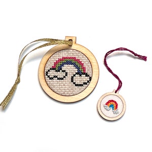 May include: Two wooden ornaments with a rainbow and clouds cross-stitch design. The larger ornament has a gold ribbon attached and the smaller ornament has a burgundy ribbon.