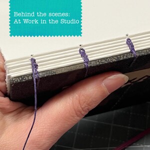 May include: A book spine being hand-bound with purple thread, showing exposed stitching through white paper signatures and a dark textured cover. A teal graphic overlay at the top contains the white text: Behind the scenes: At Work in the Studio.