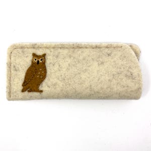 May include: A rectangular, light beige felt case featuring a brown owl appliqu&eacute; on the left side. The owl has white embroidered details on its chest, black eyes with white centers, and a black beak. The case has visible stitching along the edges.