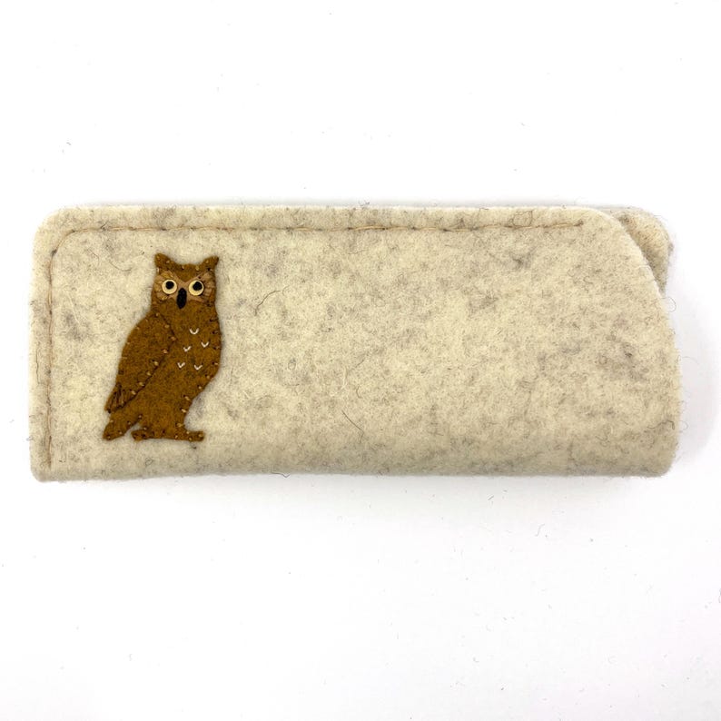 May include: A rectangular, light beige felt case featuring a brown owl appliqu&eacute; on the left side. The owl has white embroidered details on its chest, black eyes with white centers, and a black beak. The case has visible stitching along the edges.