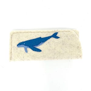 Op de afbeelding: Een rechthoekig etui van beige vilt met een applicatie van een blauwe walvis op de voorzijde. De walvis is gemaakt van twee tinten blauw vilt met zichtbare donkerblauwe stiksels langs de randen en het lichaam voor een eenvoudig, plat ontwerp.