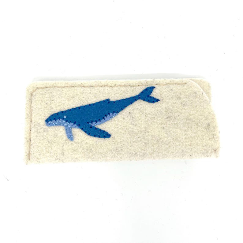 Op de afbeelding: Een rechthoekig etui van beige vilt met een applicatie van een blauwe walvis op de voorzijde. De walvis is gemaakt van twee tinten blauw vilt met zichtbare donkerblauwe stiksels langs de randen en het lichaam voor een eenvoudig, plat ontwerp.