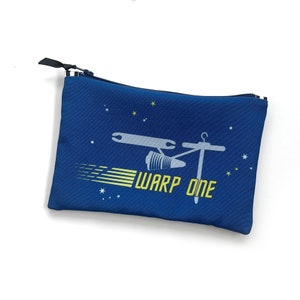 May include: A rectangular blue zippered pouch featuring a graphic of a sewing-themed spaceship. The design includes a needle and bobbin as the ship, yellow motion lines, small white and yellow stars, and the yellow text WARP ONE.