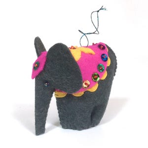 Può includere: Un ornamento di elefante in feltro grigio con una sella in feltro rosa decorata con paillettes colorate. L'elefante ha una stella gialla sulla sua sella.