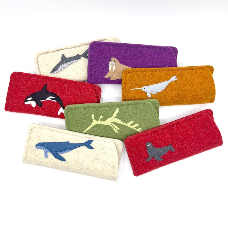 Op de afbeelding: Een collectie rechthoekige vilten etuis in diverse kleuren, elk voorzien van een opgestikte applicatie van een zeedier. De ontwerpen omvatten een haai, orka, walrus, narwal, zeedraak, walvis en zeehond op rood, paars, oranje, groen en cr&egrave;me vilt.